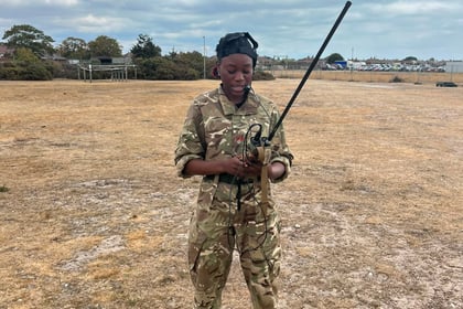 A person in camouflage military uniform stands on a dry, grassy field holding a long radio aerial during the Berkshire ACF Summer Camp 2026. The sky is partly cloudy, and there are trees and small buildings in the background.