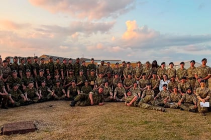 A large group of people in camouflage uniforms pose together on a grassy area outdoors at sunset during the Berkshire ACF Summer Camp 2026, with buildings and a partly cloudy sky in the background.