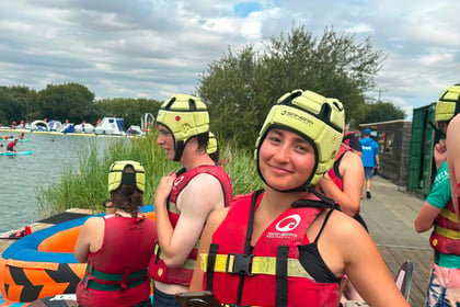 Young people wearing yellow helmets and red life jackets stand by a lake with inflatables at the Berkshire ACF Summer Camp 2026; one girl smiles at the camera while holding a mobile, others look out over the water on this cloudy day.