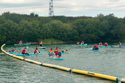 People wearing life jackets paddle and stand on blue boards in a lake, surrounded by yellow floating barriers and greenery, under a cloudy sky during the Berkshire ACF Summer Camp 2026.