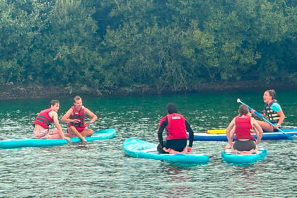 Five people wearing life jackets sit and kneel on paddleboards in a calm, greenish body of water, surrounded by dense green trees in the background—a tranquil scene from the forthcoming Berkshire ACF Summer Camp 2026.
