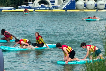 Four people in yellow life vests paddle on blue boards in a lake, racing during the Berkshire ACF Summer Camp 2026. An inflatable water park is in the background, and two others are seen further back on floating boards.