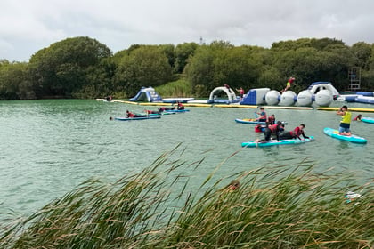 People paddle on boards and kayaks in a lake at Berkshire ACF Summer Camp 2026, wearing life jackets. An inflatable obstacle course floats in the background near green trees. Tall grass lines the shore in the foreground, and the sky is cloudy.