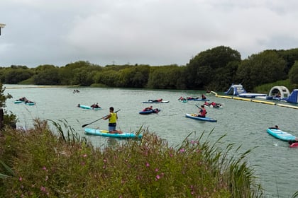 People kayaking on a river surrounded by greenery, with an inflatable obstacle course visible on the right side. Wildflowers and tall grass line the riverbank in the foreground, creating a vibrant setting for this year’s Berkshire ACF Summer Camp 2026. The sky is cloudy, adding to the adventure-filled atmosphere of the camp.