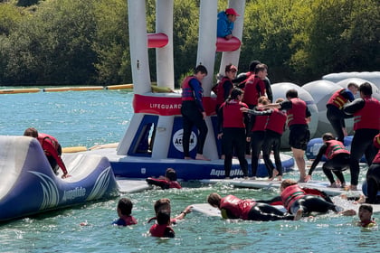 Children in red life jackets play on an inflatable water park structure in a lake at the Berkshire ACF Summer Camp 2026, some climbing, some splashing in the water, with trees and blue sky in the background. A lifeguard watches from above.