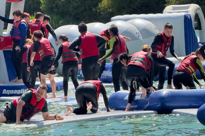 A group of people in red life jackets navigate an inflatable obstacle course on water at the Berkshire ACF Summer Camp 2026, some climbing, some standing, and some slipping, with trees in the background.