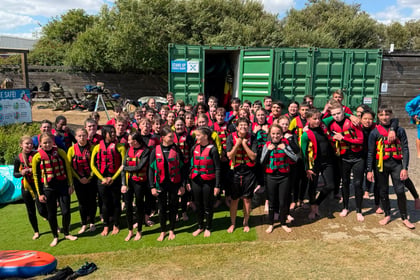 A large group of children in wetsuits and life jackets pose and smile outside on a sunny day at the Berkshire ACF Summer Camp 2026, standing on grass in front of a green shipping container and trees.