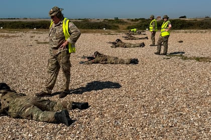 Soldiers in camouflage uniforms and high-visibility vests stand and lie on a rocky, open ground under a clear blue sky, possibly during a Berkshire ACF Summer Camp 2026 training exercise.