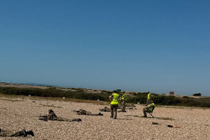 Several people in yellow vests stand and walk on a rocky field, whilst others in camouflage uniforms lie on the ground under a clear blue sky. Shrubs and flat land are visible in the background, capturing a typical training day at Berkshire ACF Summer Camp 2026.