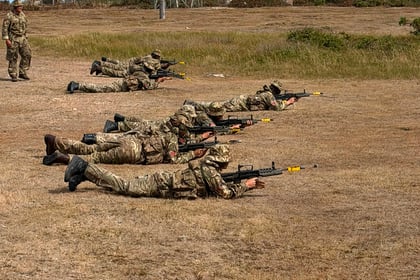 Five soldiers in camouflage uniforms lie prone on a dry field aiming rifles; a few buildings and a water tower are visible in the background under a clear sky during Berkshire ACF Summer Camp 2026.