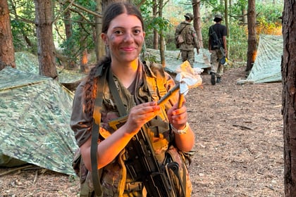 A smiling person in camouflage holds a small wrapped item in a wooded area dotted with tents at the Berkshire ACF Summer Camp 2026, whilst two others in military kit walk away in the background.