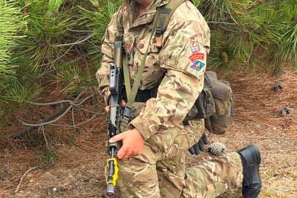 A soldier in camouflage uniform kneels outdoors with a rifle, wearing headphones and face paint, near green pine branches and dry ground during Berkshire ACF Summer Camp 2026.