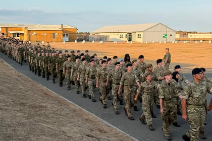 A large group of soldiers in camouflage uniforms and berets march in formation along a tarmac road, with buildings and dry grass in the background under a blue sky during the Berkshire ACF Summer Camp 2026.