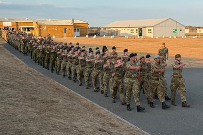 A large group of soldiers in camouflage uniforms, participating in the Berkshire ACF Summer Camp 2026, march in formation on a tarmac road near military buildings, with dry grass on either side.
