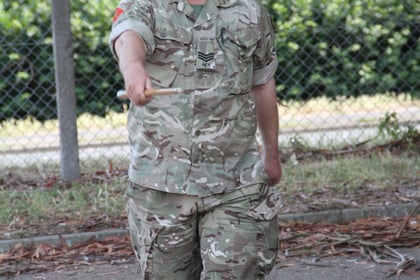 A man in camouflage military uniform walks outdoors on a paved path, smiling and making a gesture with his right hand. There are trees, a fence, and a building in the background.