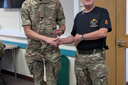 Two men in camouflage uniforms shake hands indoors. One man wears a T-shirt and glasses, while the other wears a full uniform. They stand in a classroom-like setting near a door and a wall-mounted television.