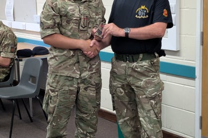 Two people in camouflage uniforms shake hands indoors, smiling. One holds a small object, possibly an award or certificate. There are empty chairs and a third person partially visible, sitting, in the background.