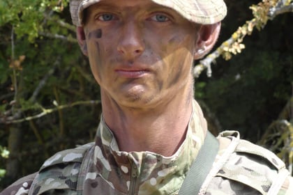 A person in camouflage military uniform and hat stands outdoors with face paint, surrounded by green foliage.