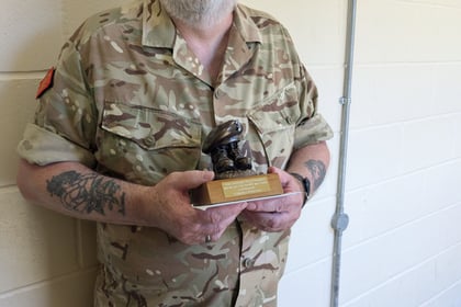 An older man in camouflage uniform and beret stands indoors, holding a small trophy. He has glasses, a white beard, and tattoos on his left forearm. The wall behind him is cream-coloured with visible wiring.