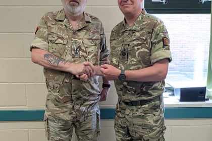 Two men in camouflage military uniforms shake hands indoors. One man wears a blue beret and glasses; the other has a tan beret. They stand in front of a white brick wall and a window.