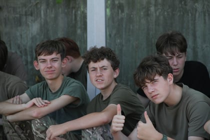 Three teenage boys in camouflage trousers and green shirts sit outdoors. Two smile at the camera; one in front gives a thumbs-up. A concrete wall is in the background, and more teenagers are partially visible.