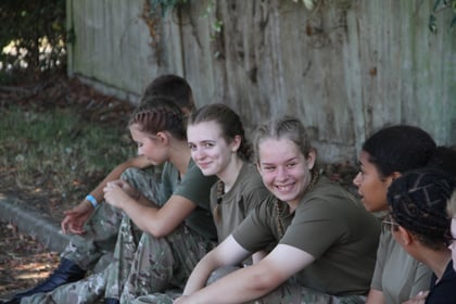 A group of young people in camouflage trousers and olive green shirts sit outdoors on the ground, smiling and talking together near a concrete wall with leafy branches above.