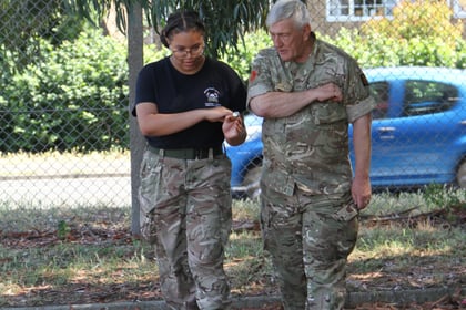 Two people in camouflage uniforms walk outside on a sunny day, looking down at their arms. A blue car and a fence are in the background, with trees providing shade.