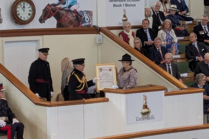 A formal ceremony at Tattersalls auction house, with a woman receiving a framed certificate from a man in uniform. Audience members are seated, and horse racing decor appears in the background.