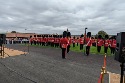 A group of guards in red uniforms and tall black hats stand in formation on a lawn, watched by a crowd behind a rope barrier under a cloudy sky.