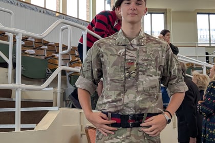 A young person in camouflage uniform and beret stands behind a table with water bottles and papers in an indoor arena with tiered seating.