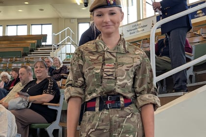 A young person in camouflage uniform and beret stands indoors, smiling. People are seated in the background on tiered seating, and one person stands near a railing. The setting appears to be a formal event or ceremony.