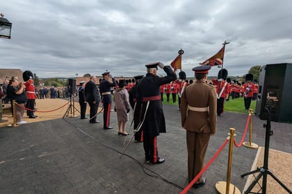 A military ceremony outdoors with soldiers in red uniforms, officers saluting, and people watching. Red ropes separate the participants from the audience under a cloudy sky.