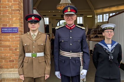 The Lord Lieutenant of Suffolk in military uniform stands between and young man in army cadet uniform and a young lady in sea cadet uniform in a doorway with seating in the background