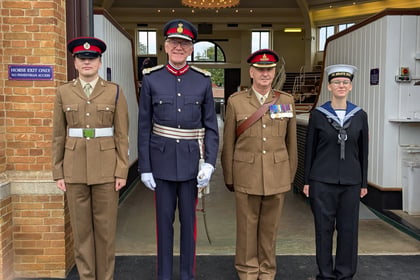 Four uniformed individuals stand side by side: two in brown army uniforms, one in a dark blue dress uniform with medals, and one in a navy uniform, all posing formally in front of a building entrance.