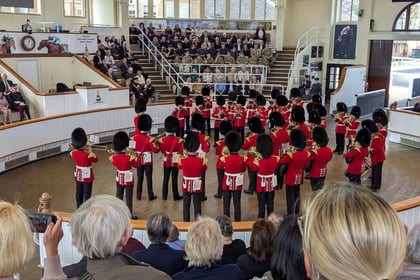 A band in red uniforms and bearskin hats performs in an indoor arena before a seated audience, with people observing and taking photos from the foreground and stands.