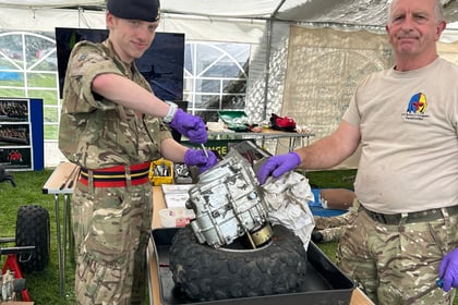Two people in military uniforms handle a metal object using purple gloves at an outdoor event, standing under a large marquee with tables and equipment around them.