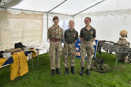 Three people in military-style uniforms stand smiling inside a white marquee, with tables displaying military kit and equipment around them. The ground is grass, and a helmet and camouflage netting are visible on the right.