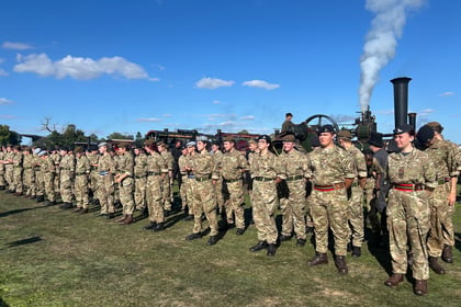 A large group of soldiers in camouflage uniforms stands in rows on a grassy field with a steam engine and blue sky in the background.