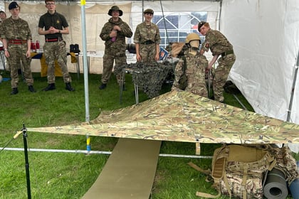 Six people in camouflage uniforms stand and sit inside a tent. Camping equipment, including a camo tarpaulin shelter, a mat, and a rucksack, is set up on grass under the tent.