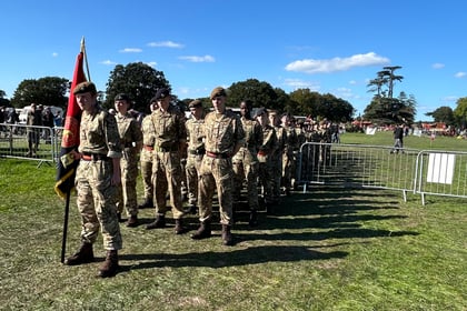 Cadets in uniform are lined up in rows of 3 into the distance. One is standing at the front with a flag. They are between two barriers waiting to march