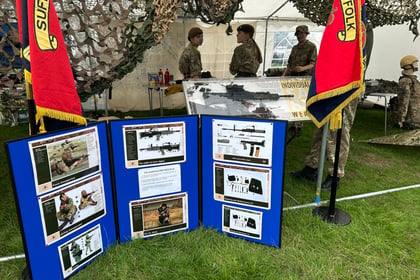 A military display marquee with soldiers, equipment, and informational boards showing images and details about weapons; Suffolk banners and camouflage netting are visible.
