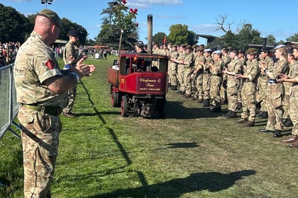 A group of soldiers in uniform stand in formation outdoors, applauding a small vintage steam vehicle with a red front. Blue sky and grass field are visible.