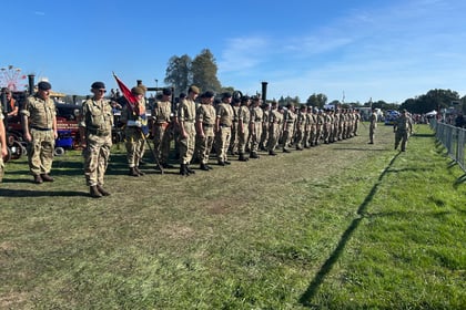 A line of soldiers in camouflage uniforms and berets stands to attention on a grassy field during an outdoor event, with people and vehicles in the background under a clear blue sky.
