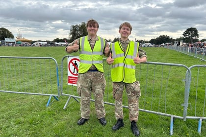 Two young people in camouflage uniforms and yellow vests stand by a metal barrier on grass, both giving thumbs up. A no entry sign is on the barrier, and there's a crowd and trees in the background under a cloudy sky.