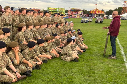 A group of young people in camouflage uniforms and berets sit on grass, listening to a man standing in front of them. Funfair rides and vintage cars are visible in the background.
