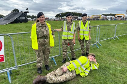 Four people in camouflage uniforms and high-visibility vests stand and pose by a metal barrier at an outdoor event. One person is lying on the grass in front, smiling. Crowds and marquees are in the background under a cloudy sky.