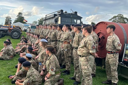 A group of uniformed cadets stand and sit in formation on grass near military vehicles, outdoors under a cloudy sky.
