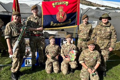 Seven people in camouflage uniforms stand and kneel on grass, posing by a Suffolk Regiment flag and tents on a sunny day. Some hold items; a blue sky with clouds is visible above.