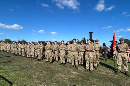 A large group of soldiers in camouflage uniforms stand in formation on grass under a blue sky, with a flag and old train in the background.