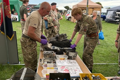 Two people in military uniforms work on machinery at a table filled with tools under a marquee. Others in uniform and marquees are visible in the background on a grassy field.
